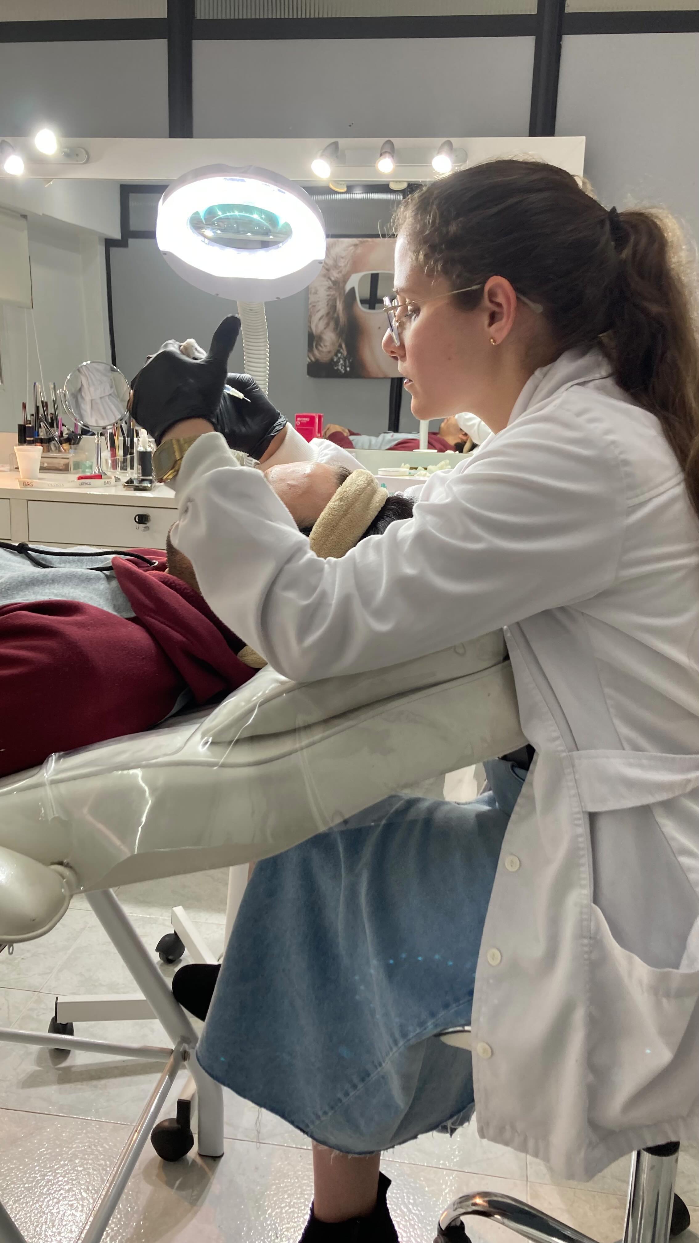 Una profesional de Bella con guardapolvo y guantes aplica un tratamiento a un cliente recostado en la camilla, bajo una lámpara de aumento
